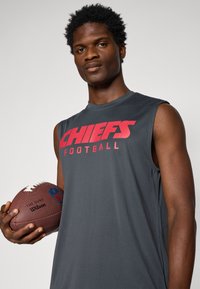 Bežila pilka beisbolo marškiniai pilkos spalvos be rankovių su užrašu "CHIEFS FOOTBALL" ryškiu raudonu šriftu. Modelis laiko rudą futbolą su baltais raiščiais ir NFL logotipu.