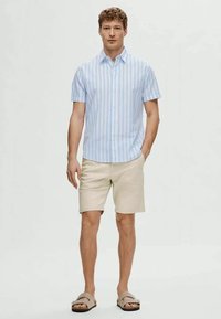 Chemise à manches courtes rayée bleu clair et blanche, shorts beiges et sandales. La chemise présente un design à boutons et une coupe décontractée.