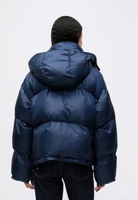 Námořnická modrá péřová bunda s velkou kapucí, lesklým povrchem a prošívaným designem. Disponuje volným střihem a viditelným švem.