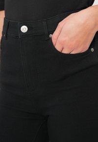 Main dans la poche avant d'un jean noir ajusté, avec des boutons et des rivets en argent visibles.