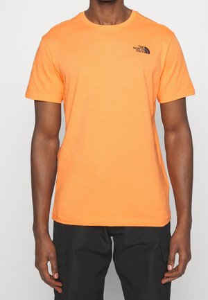 Mężczyzna w jasnopomarańczowej koszulce z krótkim rękawem z logo The North Face na piersi oraz czarnych spodniach typu cargo na jednolitym tle.