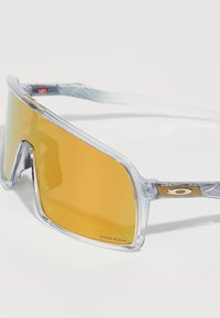 Óculos de sol com uma armação de plástico transparente e grandes lentes amarelas, apresentando um detalhe dourado nas hastes. Lentes rotuladas "PRIZM."