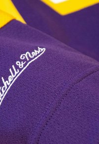Lila Stoff mit einer strukturierten Oberfläche, ausgestattet mit einem weißen gestickten Logo "Mitchell & Ness" am Ärmel, akzentuiert durch einen gelben Kragen.