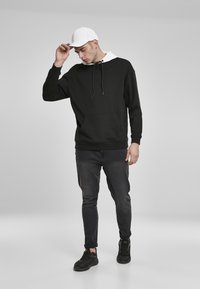 Fekete kapucnis pulóver fehér díszítésekkel, zsinórral és elülső zsebbel; sötétszürke farmernadrággal és fekete sportcipővel párosítva, fehér sapkával a fején.