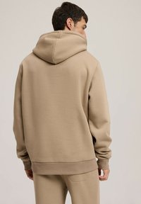 Beżowy kaptur wykonany z miękkiej tkaniny, cechujący się luźnym krojem, ściągaczami na rękawach oraz kieszenią typu kangur. Widok z tyłu podkreśla bezszwowy design.