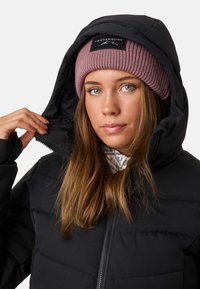 Zwarte pufferjack met rits, capuchon en geometrisch geborduurd patroon. Roze ribgebreide beanie met een zwart logo patch. Haar lichtjes getextureerd.