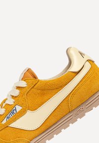 Sneaker in suede giallo con dettagli crema, lacci bianchi e finitura testurizzata. Presenta un logo sul lato e una suola in gomma marrone chiaro.