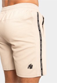 Beige sportieve shorts van een textuurstof, met een elastische tailleband, zijzak en een zwart logo aan de onderkant links. Een zwarte zijstrik met de tekst "GORILLA WEAR."