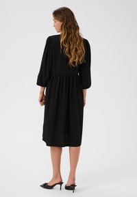 Black midi dress with puffed sleeves, fitted bodice, and gathered waist, featuring a smooth texture. Worn with black mules, back view shown.