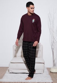 Burgundi hosszú ujjú ing karaktermintával és fekete kockás pizsama nadrág piros és fehér részletekkel, viselve egy álló férfi által.