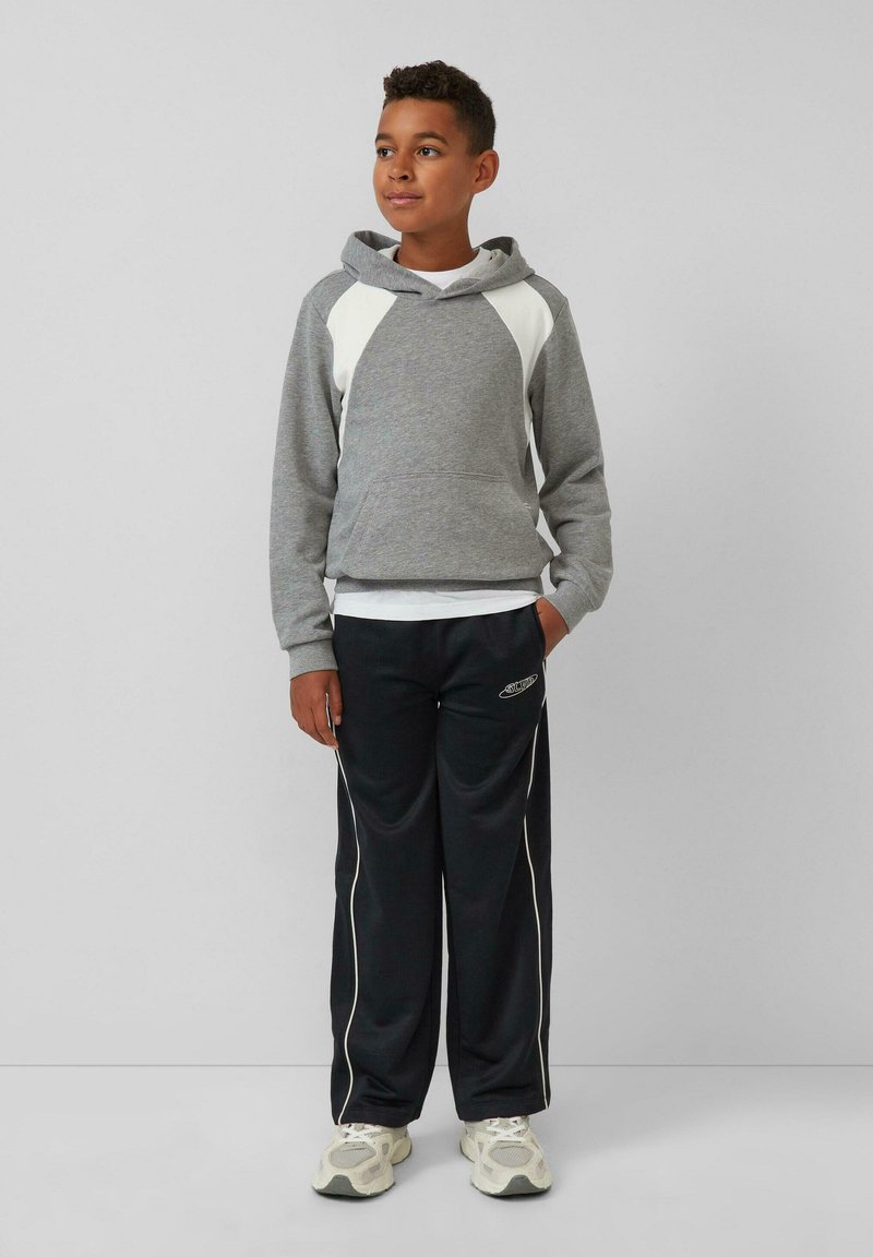 Garçon debout portant un sweat à capuche gris avec des panneaux blancs, un pantalon de survêtement noir avec des bandes blanches, et des baskets blanches, regardant vers la droite.
