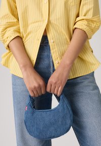 Bolsa de ganga com forma arredondada, cor azul e costura visível. Usada na mão, combinada com uma camisa amarela riscada e jeans azul claro.