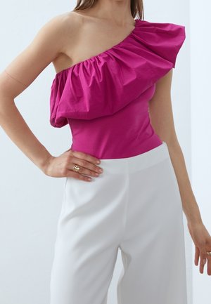 Kobieta w jasnoróżowej, asymetrycznej bluzce z falbanami, zestawionej z białymi, szerokimi spodniami z wysokim stanem, ręka na biodrze.