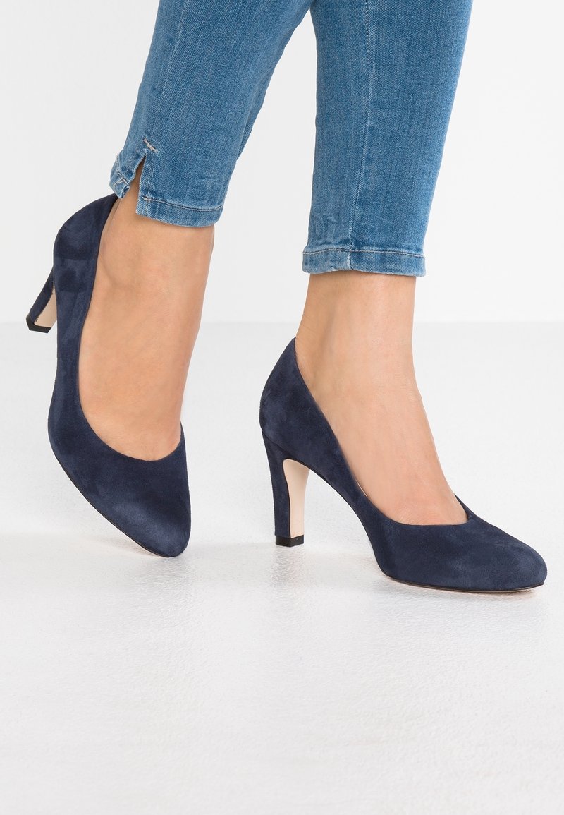 Pieds portant des chaussures à talons hauts en daim bleu associées à un jean en denim bleu avec de petites fentes à la cheville sur un sol blanc.