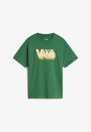 Zielona koszulka z krótkim rękawem z białym i żółtym logo "VANS", zestawiona z czarnymi spodniami i czarnymi trampkami z białymi sznurowadłami. Sweter zawiązany w talii.