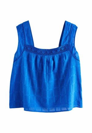 Kvinde i blå ærmeløs top og lysegrå bukser, stående på en træveranda med udsigt over havet og grøntområde.