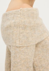 Beige, gebreide trui met een geribbelde textuur, off-the-shoulder ontwerp en een omgevouwen kraag, die de zachte, warme stof benadrukt.