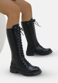 Botas de cuero negro hasta la rodilla con un cierre de cordones en la parte frontal, cremallera lateral y suela de goma resistente con un ligero tacón. Acabado texturizado en el material.