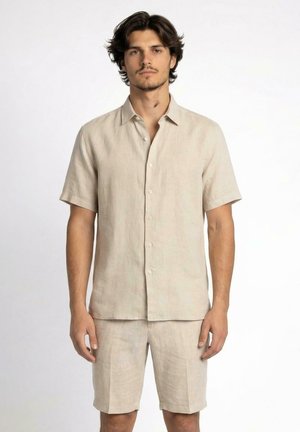 Giovane uomo con capelli scuri che indossa una camicia beige a maniche corte con bottoni e pantaloncini beige abbinati, in piedi davanti a uno sfondo semplice.