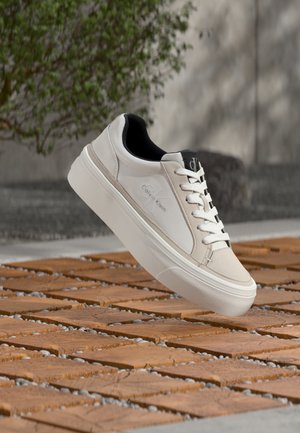 Biely teniska Calvin Klein s čiernym vnútorným lemom a bielymi šnúrkami vznášajúca sa nad dlaždičkovou drevenou a štrkovou vonkajšou plochou.