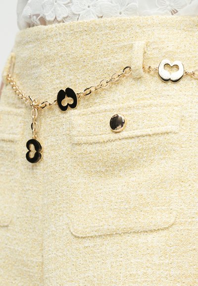 Ceinture chaîne en or avec des maillons en forme de cœur noirs et dorés portée sur une jupe texturée jaune pâle avec des poches à rabat et un bouton doré.