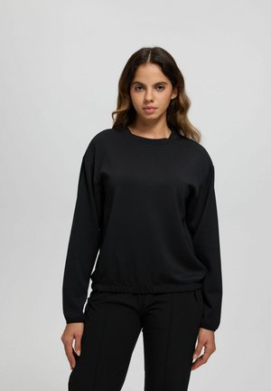 Donna con capelli ondulati che indossa una felpa nera a maniche lunghe semplice e pantaloni neri, in piedi davanti a uno sfondo grigio chiaro uniforme.