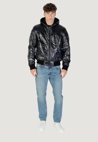 Giubbotto a vento nero lucido con zip frontale e polsini a coste, abbinato a jeans azzurri chiari e sneakers bianche.