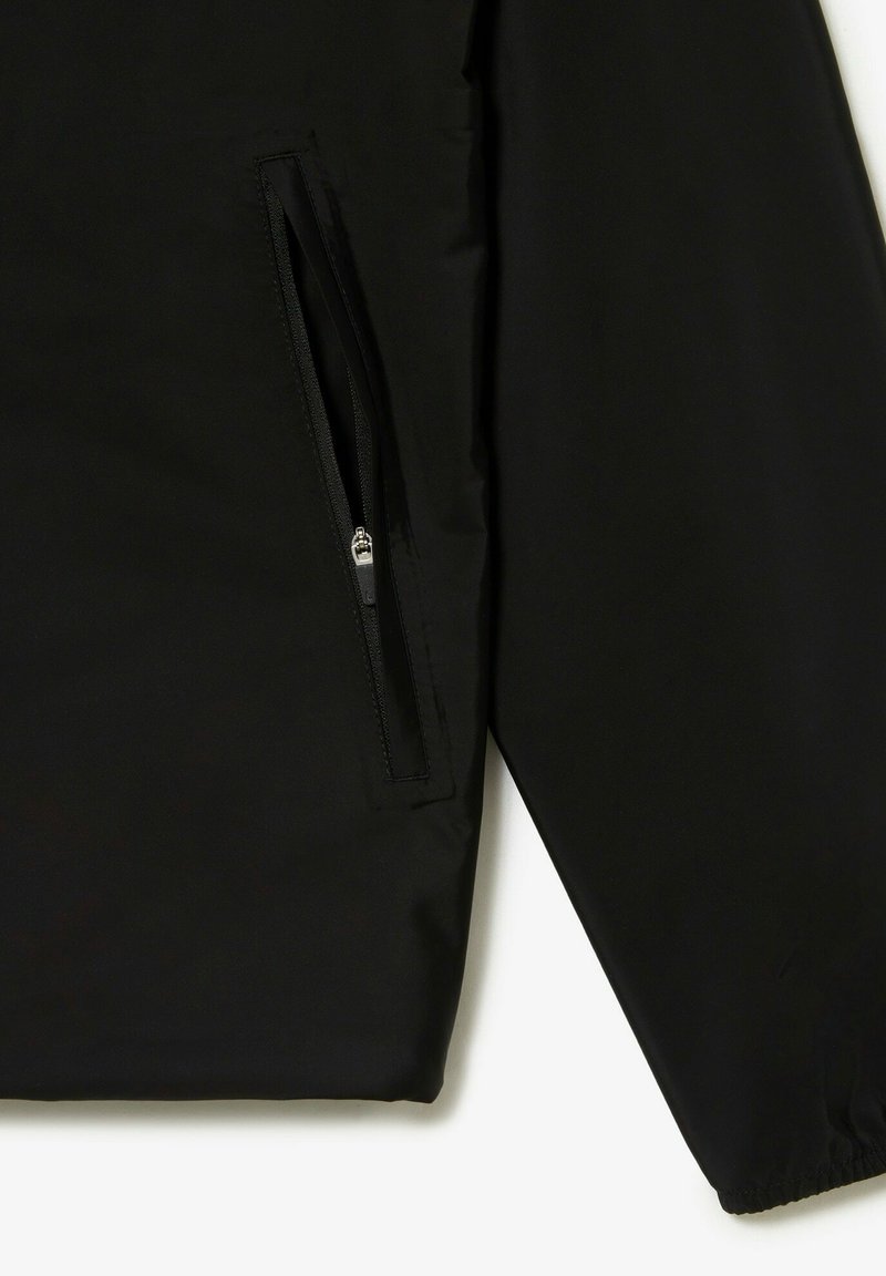 Veste coupe-vent noire avec une poche zippée et des poignets élastiques. Tissu lisse avec un léger éclat et des détails de couture minimaux.