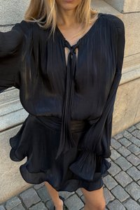 Melnā, drapētā blūze ar saites detaļu pie kakla, platām piedurknēm un pieguļošu jostu. Mīksta, raksturīga auduma struktūra ar plūstošu krišanu.