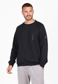 Sudadera negra con mangas raglán, tejido texturado, un bolsillo en el pecho con cremallera y un dobladillo acanalado. Combinada con pantalones gris claro.