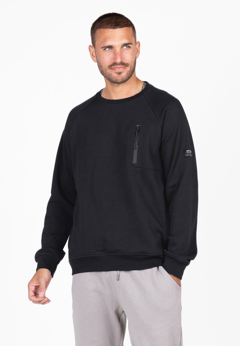 Sudadera negra con mangas raglán, tejido texturado, un bolsillo en el pecho con cremallera y un dobladillo acanalado. Combinada con pantalones gris claro.