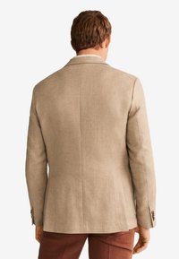Muž v béžovém saku a hnědých kalhotách, zobrazený zezadu na bílém pozadí.