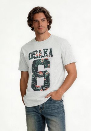 Junger Mann mit dunklen Haaren trägt ein hellgraues T-Shirt mit „OSAKA“ und floralem Nummer-6-Design, kombiniert mit blauen Jeans, stehend vor weißem Hintergrund.