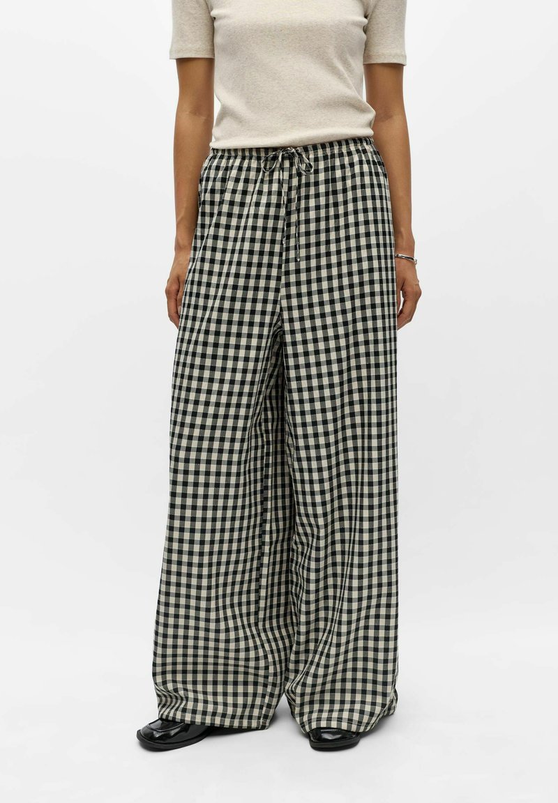 Pantalon large fluide à carreaux noir et blanc avec taille à cordon, porté avec un haut beige clair à manches courtes et des chaussures noires.