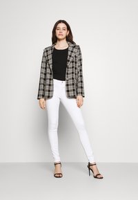 Blazer xadrez em preto e branco com lapelas caneladas, combinado com jeans brancos ajustados e saltos pretos com tiras, contra um fundo simples.