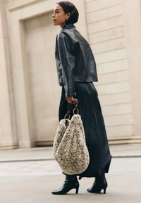 Veste courte en cuir noir, longue jupe noire en satin, et bottines noires à talons pointues. Tenant un grand sac en fausse fourrure beige avec des accents marron.