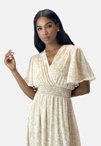 Robe florale beige avec des manches bouffantes, un décolleté en V, une taille smockée et une jupe fluide. Tissu léger avec des motifs de fleurs blanches.