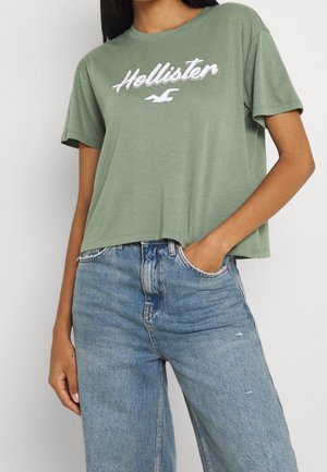 Zelené bavlněné tričko s cropped designe, které má bílé vyšití loga "Hollister" a krátké rukávy, kombinované s vysokými modrými džíny.