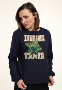 Felpa blu navy con un vivace grafico di un dinosauro e il testo "Domatore di Dinosauri". Maniche lunghe, polsini a costine e una consistenza morbida.