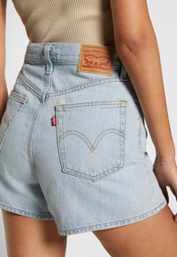 Pantalones cortos de mezclilla azul claro con cintura alta, que cuentan con una etiqueta de cuero de marca y detalles de costura visibles en los bolsillos traseros.