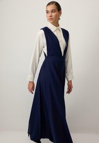 Rochie bleumarin cu decolteu adânc în V, purtată peste o cămașă albă cu nasturi. Rochia are pliuri și o talie ajustată, creând o siluetă fluidă.