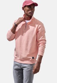 Light pink zip-neck sweatshirt with ribbed cuffs and hem. Worn over a white shirt, paired with light grey jeans. Patch logo at the hem.