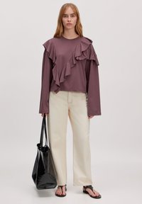 Lila langärmeliges Oberteil mit Rüschendetails, kombiniert mit hellen, weit geschnittenen Hosen und einer schwarzen Handtasche. Flache Sandalen runden den Look ab.