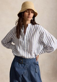 Kvinne med langt mørkt hår som har på seg en beige bøttehatt, hvit bluse med blå, vertikale striper og blå bukser med høy midje og belte.