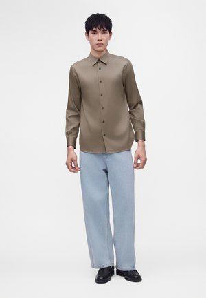 Ung mand står iført en brun knap-op langærmet skjorte, lyseblå jeans med vide ben og sorte sko mod en ensfarvet hvid baggrund.