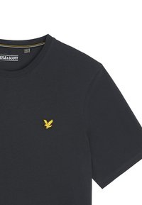 T-shirt di cotone blu navy con scollatura rotonda, maniche corte e piccolo logo giallo a forma di aquila sul lato sinistro del petto. Vestibilità standard e design semplice.