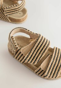 Beige vävda sandaler med svarta ränder, med två justerbara kardborreband, en mjuk insida och en strukturerad, lätt sula.