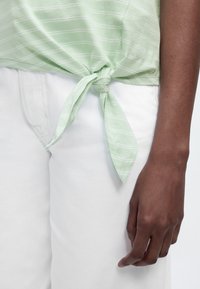 Άτομο που φοράει λευκό παντελόνι και ανοιχτό πράσινο ριγέ μπλουζάκι δεμένο με κόμπο στη μέση, με δεξί χέρι ορατό να ακουμπάει δίπλα στο ισχίο.