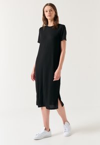 Schwarzes knielanges Kleid mit kurzen Ärmeln, seitlichen Schlitzen, gerippter Textur und lockerer Passform. Kombiniert mit weißen Sneakern.