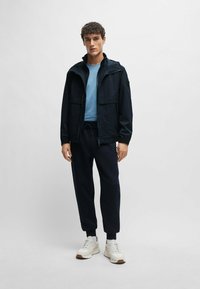Giacca impermeabile blu navy con chiusura a zip, due tasche frontali, abbinata a una t-shirt azzurro chiaro e pantaloni della tuta scuri, completata da sneakers bianche.